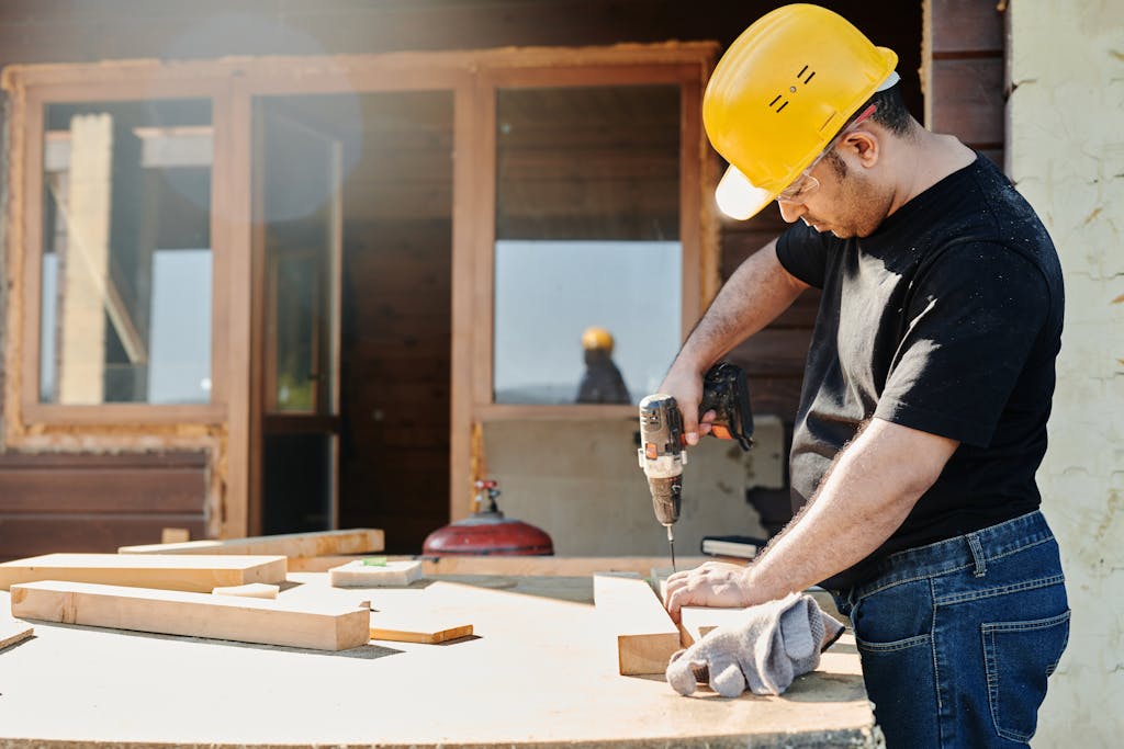 A construction worker with a hard hat uses a power drill on a wooden plank at a renovation site.
